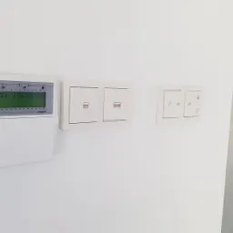 Panel sterowania alarmem z wyświetlaczem LCD pokazującym datę i godzinę, obok dwa podwójne włączniki z ikonami bram oraz pojedynczy włącznik z ikonami młotka, samochodu i domu, zamontowane...