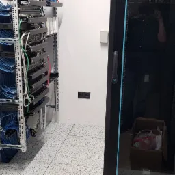 Szafa serwerowa z widocznymi kablami sieciowymi w pomieszczeniu z szarą podłogą, obok ściana z gniazdami elektrycznymi i biała szafka.