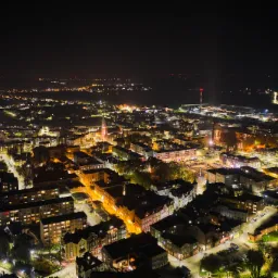Nocny widok z lotu ptaka na oświetlone miasto z dominującą wieżą kościoła, w tle widoczne światła portu i odległe wzgórza.
