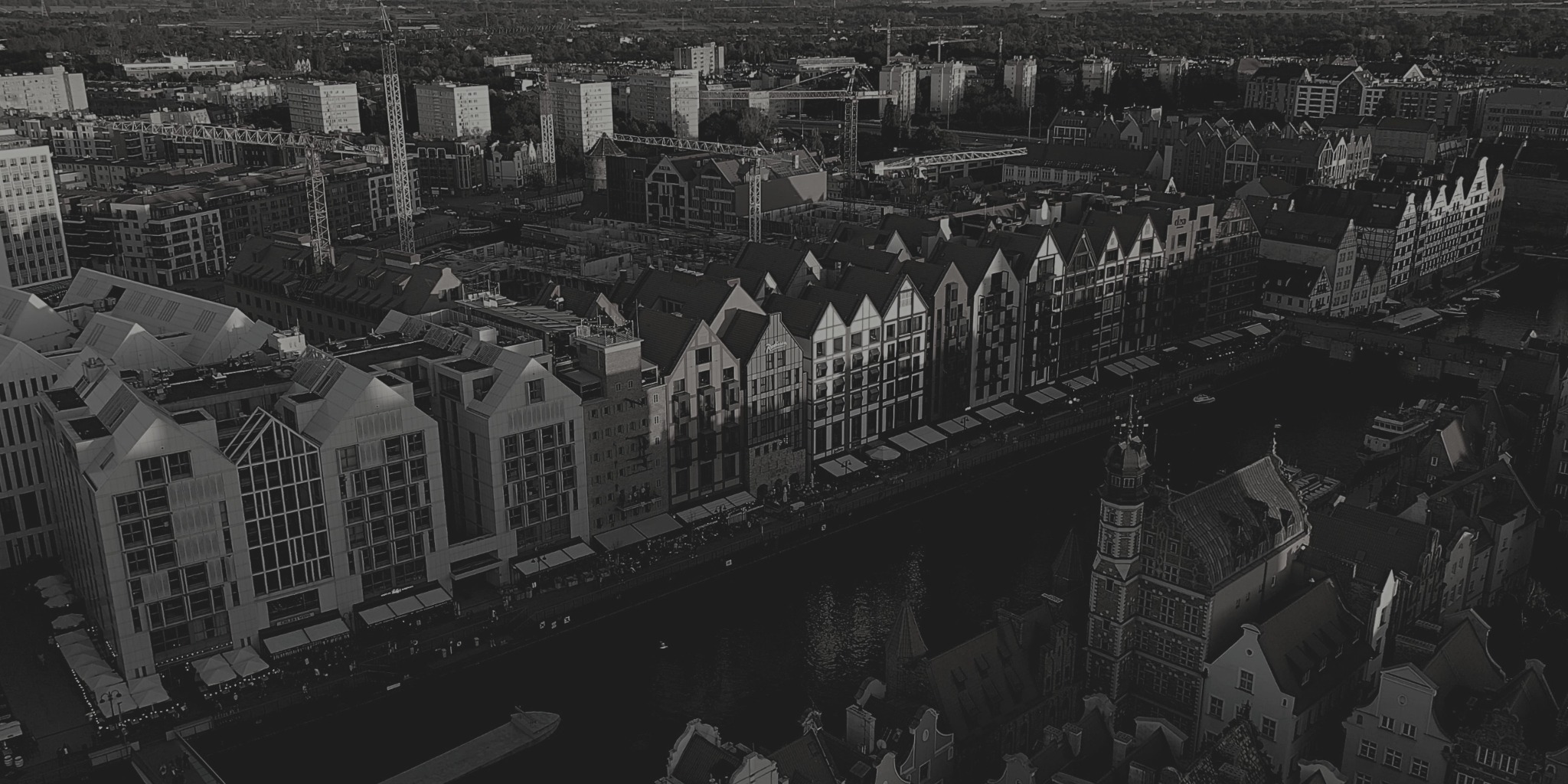 Czarno-białe ujęcie z lotu ptaka na gdańską starówkę z rzeką Motławą, widoczne charakterystyczne kamienice i żurawie budowlane w tle.