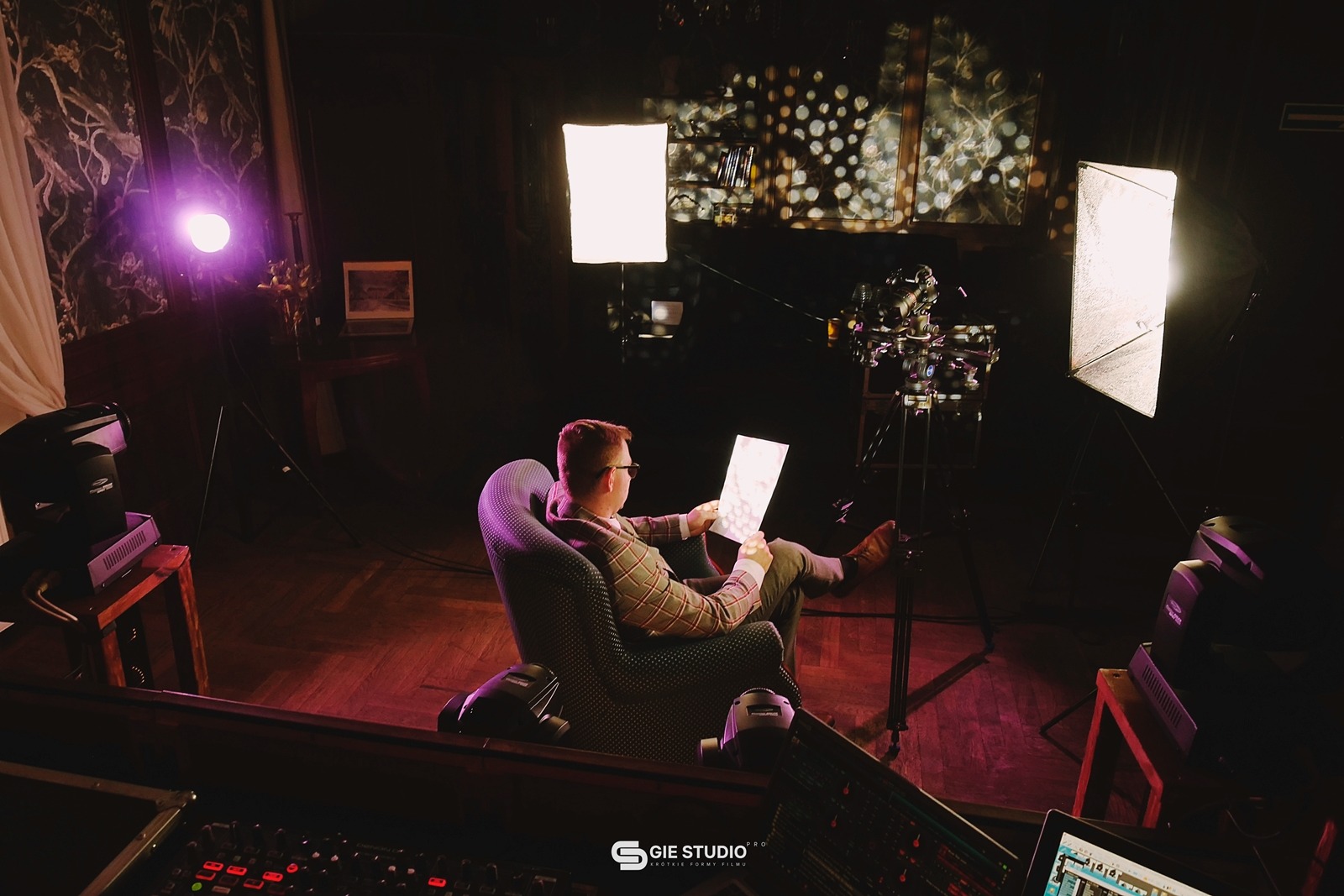 Mężczyzna w fotelu przegląda storyboard na planie filmowym z oświetleniem i kamerą na statywie, widoczny stół mikserski z laptopem.