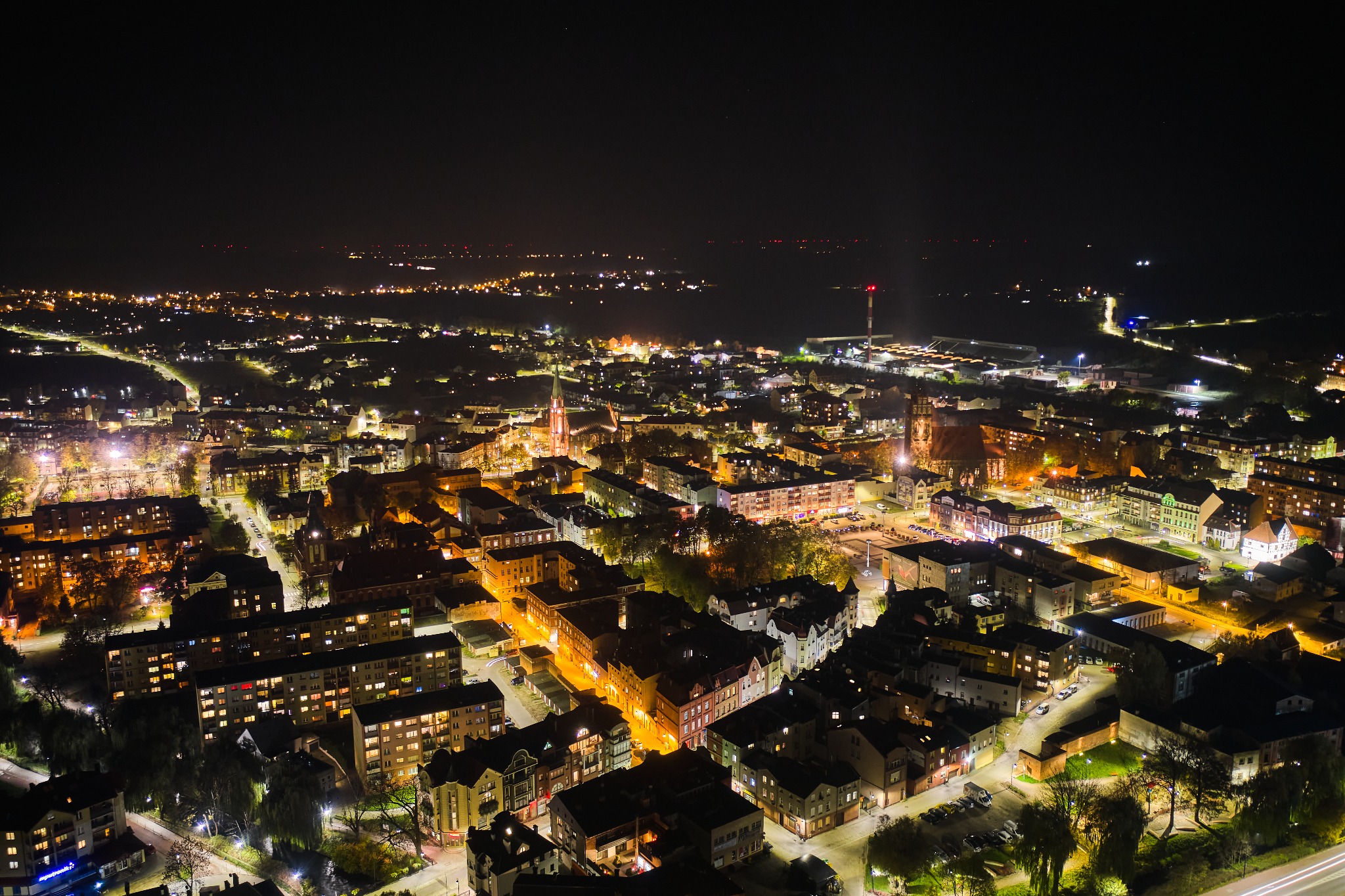 Nocny widok z lotu ptaka na oświetlone miasto z dominującą wieżą kościoła, w tle widoczne światła portu i odległe wzgórza.