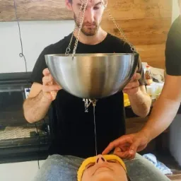 Mężczyzna wykonuje zabieg polegający na polewaniu czoła pacjentki ciepłym płynem z metalowej misy zawieszonej na łańcuchach, podczas sesji ajurwedyjskiej.