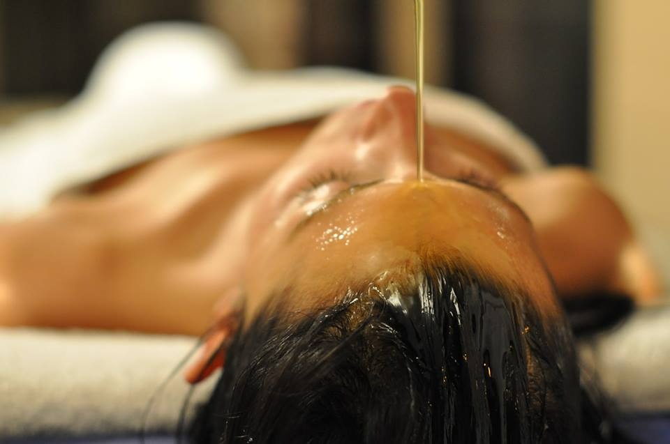 Kobieta leżąca na plecach z zamkniętymi oczami, na jej czoło spływa cienki strumień olejku podczas zabiegu relaksacyjnego.