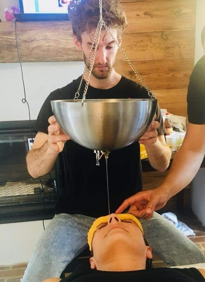 Mężczyzna wykonuje zabieg polegający na polewaniu czoła pacjentki ciepłym płynem z metalowej misy zawieszonej na łańcuchach, podczas sesji ajurwedyjskiej.