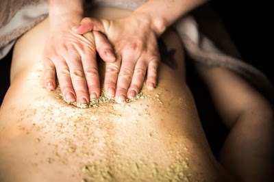 Dłonie nakładają peeling z drobinkami na plecy klienta leżącego na stole do zabiegów, widoczny fragment tatuażu na boku.