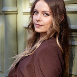 Paweł Kadruje - Fotografia - Portret kobiety w brązowym żakiecie, opierającej się o jasnozielone drzwi z kolumnami. Uśmiech, długie włosy z pasemkami, makijaż podkreślający oczy.