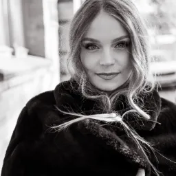 Czarno-biały portret kobiety o jasnych włosach, w ciemnym futrze, z subtelnym uśmiechem i promieniejącym spojrzeniem, na rozmytym tle miejskim.