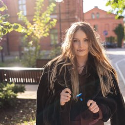 Paweł Kadruje - Fotografia - Portret kobiety w brązowym garniturze i futrze na tle ceglanego budynku, oświetlonego promieniami słońca. Ujęcie z dołu podkreśla architekturę i jesienny klimat.