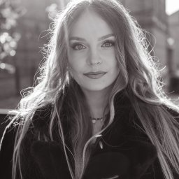 Paweł Kadruje - Fotografia - Czarno-biały portret kobiety z długimi włosami, patrzącej w obiektyw. Delikatne światło słoneczne podkreśla rysy twarzy. W tle widoczna latarnia i fragment budynku.