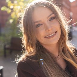 Paweł Kadruje - Fotografia - Portret uśmiechniętej kobiety w brązowej marynarce, z promieniami słońca we włosach, wykonany w plenerze w Łodzi. Ujęcie z bliska z delikatnym rozmyciem tła.