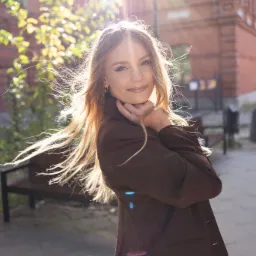 Uśmiechnięta blondynka w brązowym żakiecie pozuje na tle ceglanego budynku i zieleni w słoneczny dzień. Włosy rozwiane wiatrem, delikatny makijaż.