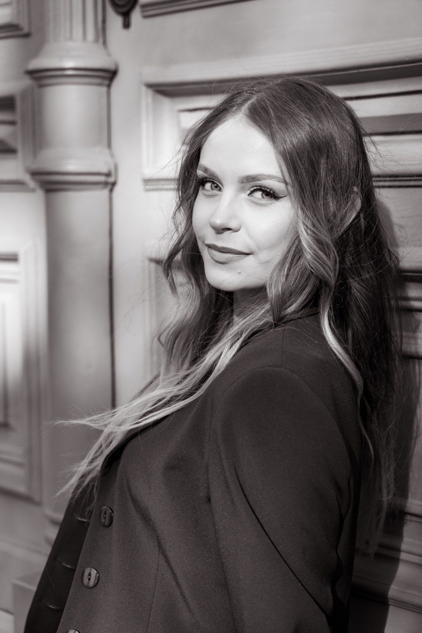 Portret kobiety w czarno-białej tonacji, z długimi, falowanymi włosami, opierającej się o ścianę z dekoracyjnymi panelami. Delikatny uśmiech, profesjonalny makijaż.
