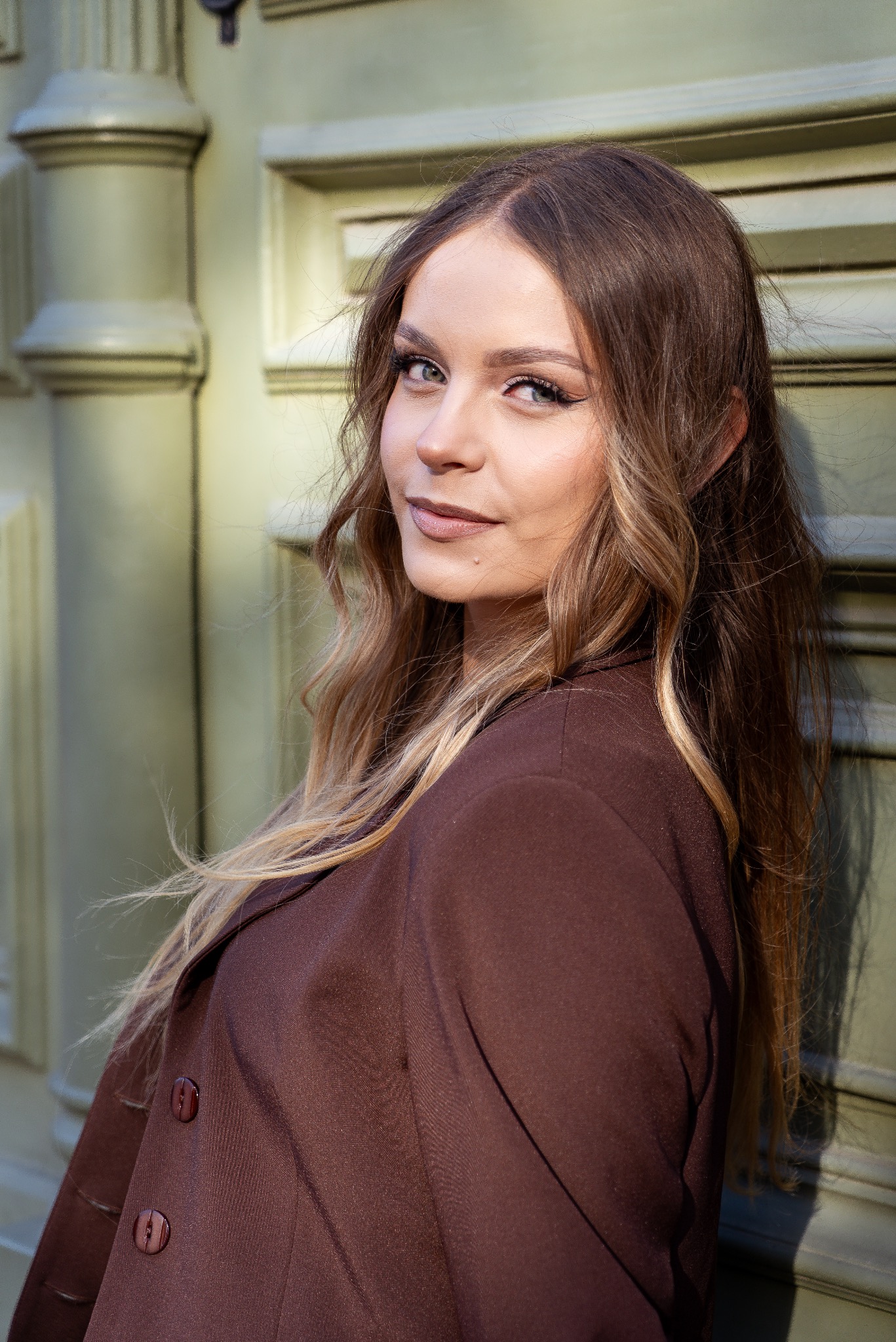 Portret kobiety w brązowym żakiecie, opierającej się o jasnozielone drzwi z kolumnami. Uśmiech, długie włosy z pasemkami, makijaż podkreślający oczy.