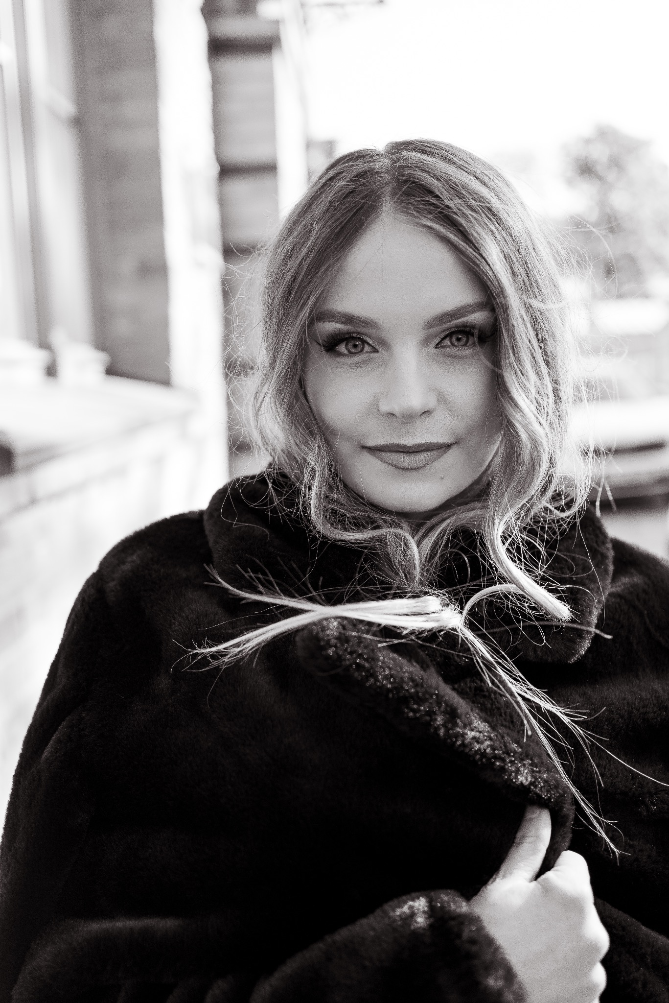 Czarno-biały portret kobiety o jasnych włosach, w ciemnym futrze, z subtelnym uśmiechem i promieniejącym spojrzeniem, na rozmytym tle miejskim.