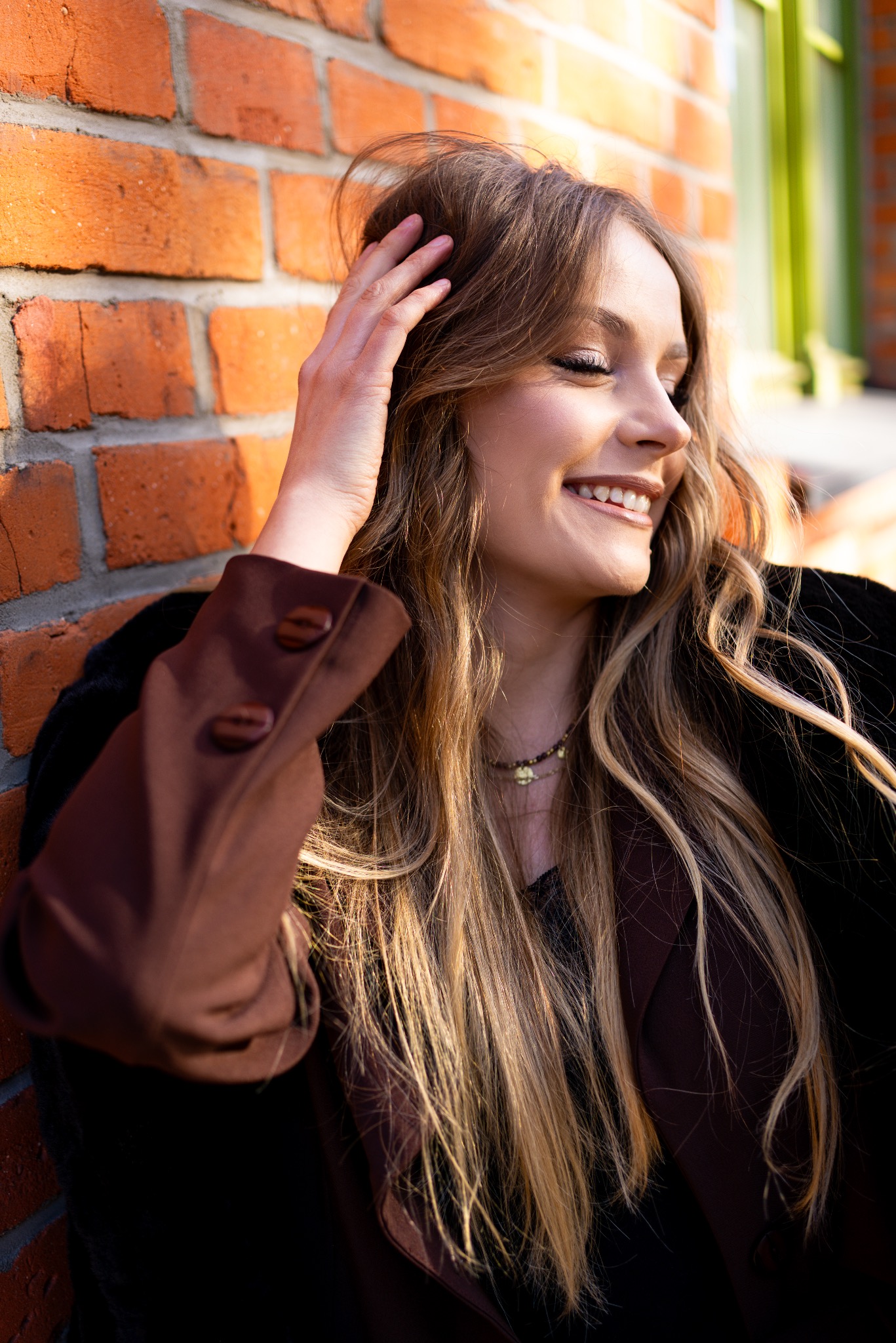 Uśmiechnięta blondynka opiera się o ceglaną ścianę, dłonią poprawiając włosy. Na szyi delikatny naszyjnik, ubrana w ciemną bluzkę i brązową marynarkę. Ciepłe światło słoneczne.