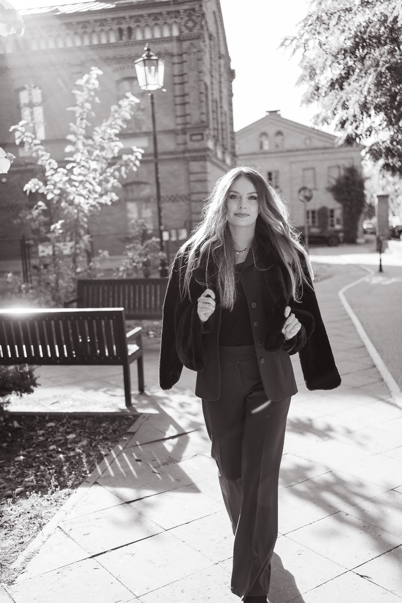 Czarno-białe zdjęcie kobiety w garniturze i futrze na tle ulicy z budynkami i drzewami, oświetlonej promieniami słońca.