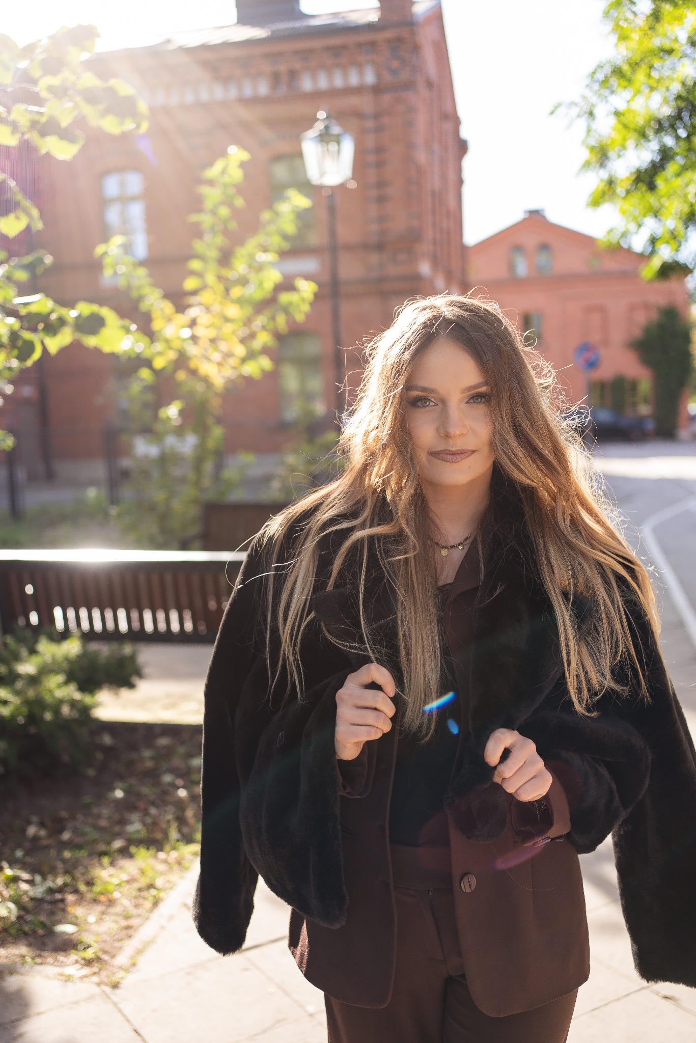 Portret kobiety w brązowym garniturze i futrze na tle ceglanego budynku, oświetlonego promieniami słońca. Ujęcie z dołu podkreśla architekturę i jesienny klimat.