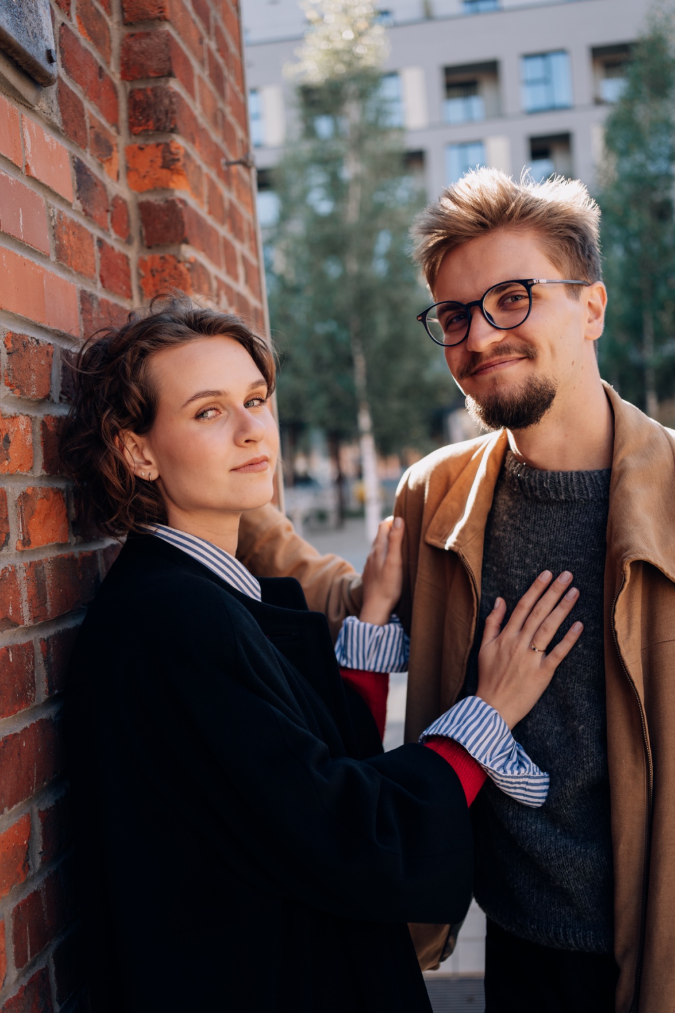 Uśmiechnięta para w okularach pozuje na tle ceglanej ściany i nowoczesnego budynku. Kobieta opiera się o ścianę, a mężczyzna obejmuje ją ręką. Naturalne światło.