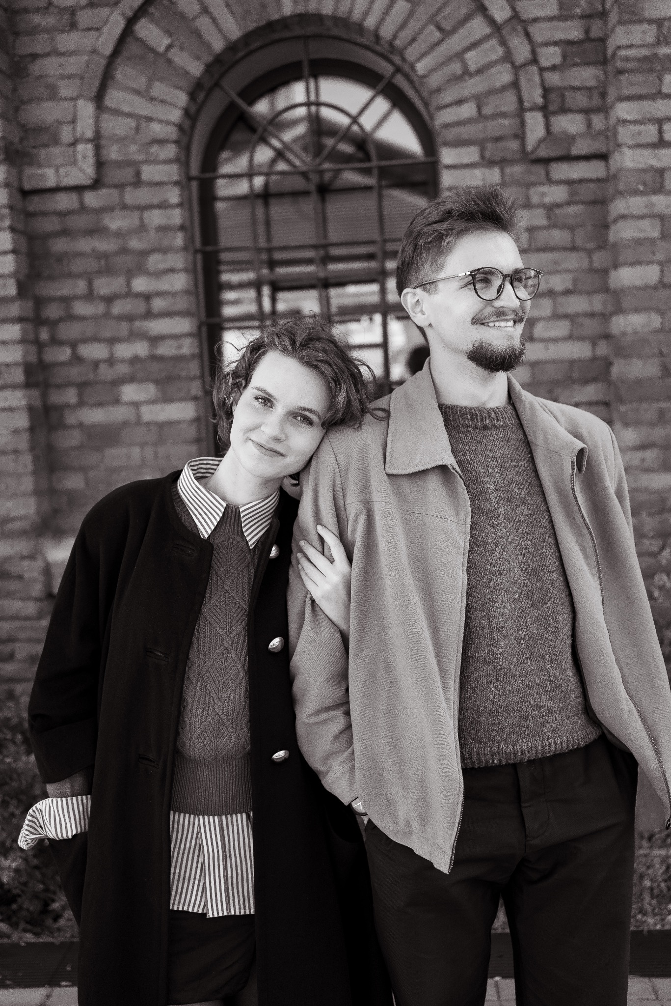 Czarno-białe zdjęcie portretowe pary, kobieta opiera głowę na ramieniu mężczyzny. Ceglane tło z oknem w tle dodaje charakteru. Ubrani w swetry i płaszcze.