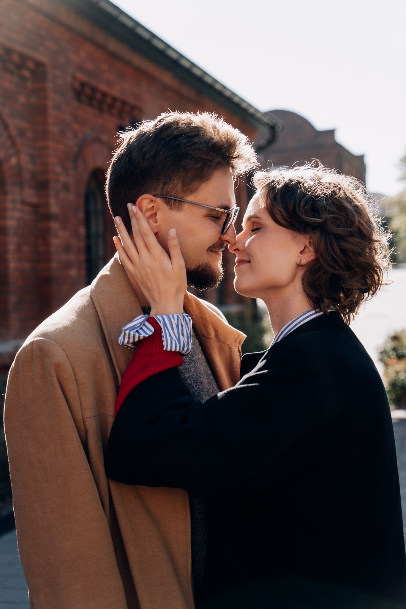 Zakochana para w zbliżeniu, dotykająca się nosami na tle ceglanego budynku. Kobieta w czarnej marynarce, mężczyzna w beżowym płaszczu i okularach.
