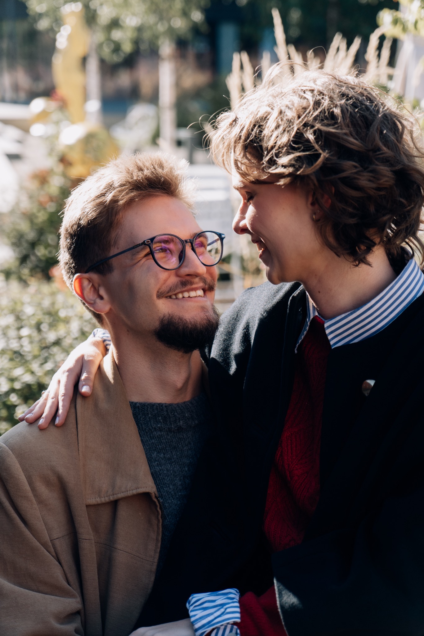 Uśmiechnięta para patrzy na siebie w słoneczny dzień. Mężczyzna w okularach i beżowej kurtce, kobieta w ciemnej marynarce i koszuli w paski. Romantyczna atmosfera.
