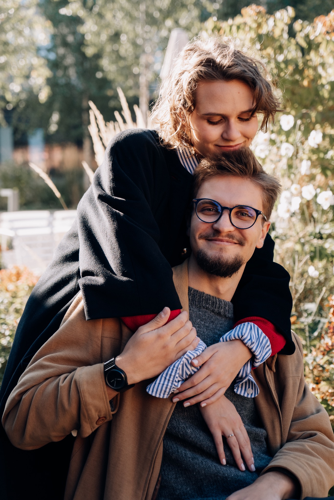 Uśmiechnięta para w jesiennej scenerii: kobieta obejmuje mężczyznę w okularach, oboje ubrani w stylowe płaszcze na tle kwitnących krzewów i drzew.