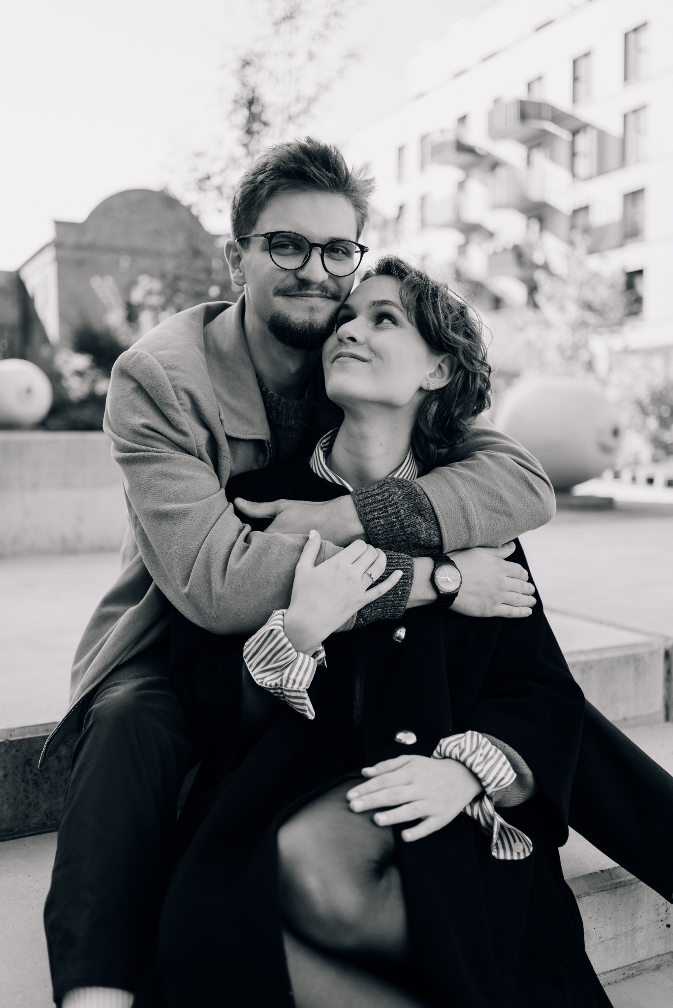 Czarno-biała fotografia zakochanej pary siedzącej na schodach. Mężczyzna w okularach obejmuje kobietę, która spogląda na niego z uśmiechem. W tle budynek.