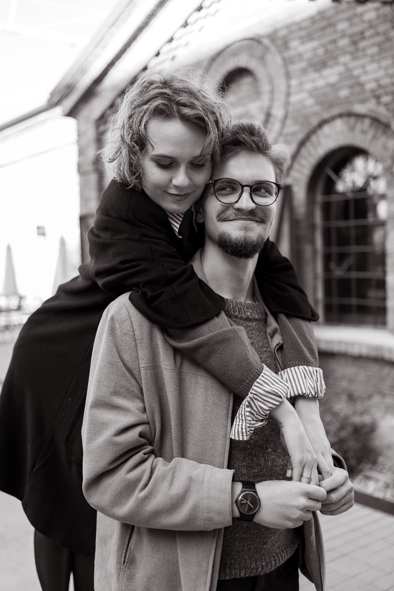 Czarno-białe zdjęcie uśmiechniętej pary: kobieta obejmuje mężczyznę od tyłu na tle ceglanej ściany z łukowatymi oknami. Ona patrzy w dół, on w górę. Widoczny zegarek na ręce.