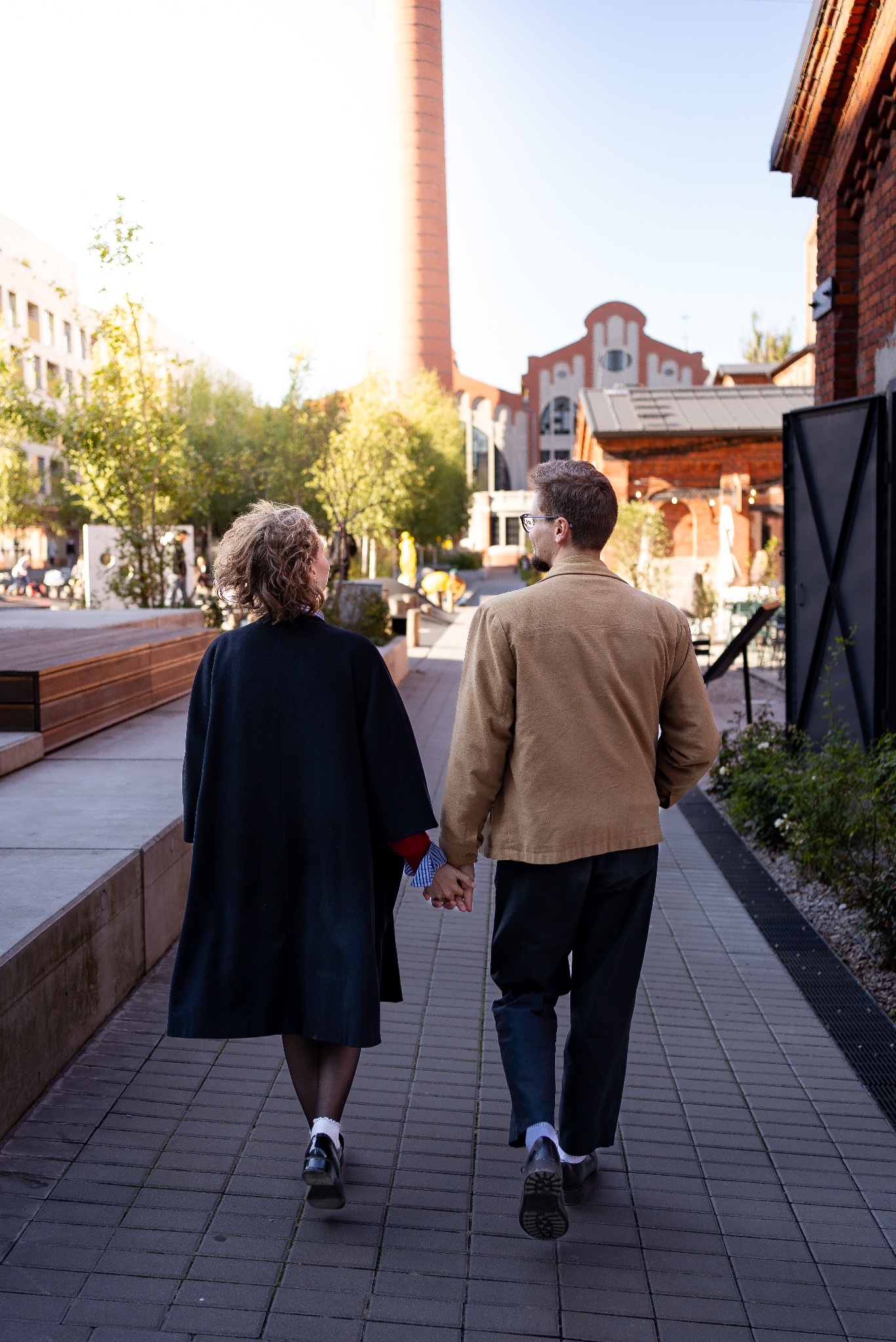 Para spaceruje trzymając się za ręce w otoczeniu ceglanej architektury i wysokiego komina. Ujęcie z tyłu, w słoneczny dzień, na tle miejskiego krajobrazu.
