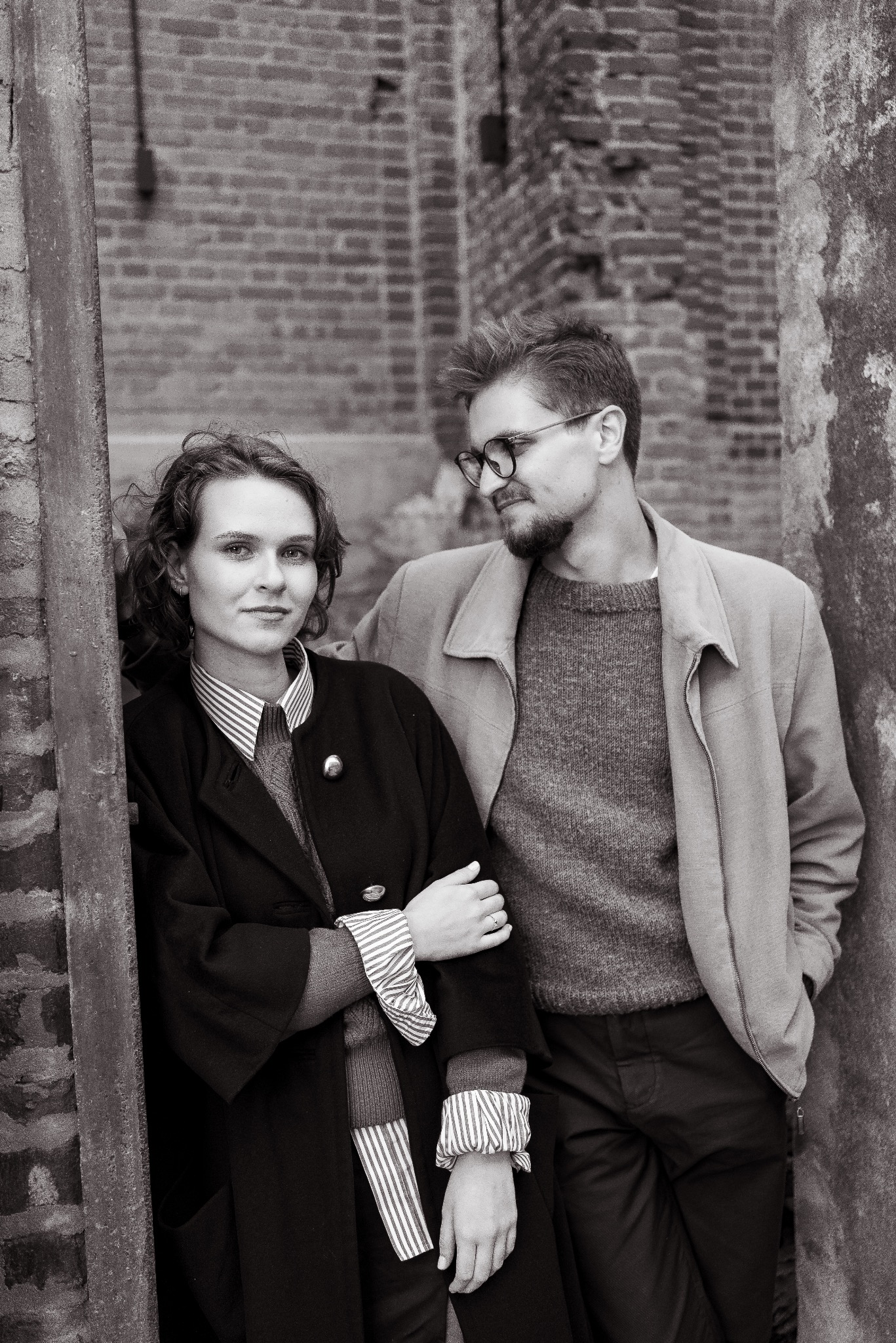 Czarno-biała fotografia portretowa pary w objęciach, opartych o ceglaną ścianę. Kobieta patrzy w obiektyw, mężczyzna spogląda w bok, oboje w eleganckich strojach.