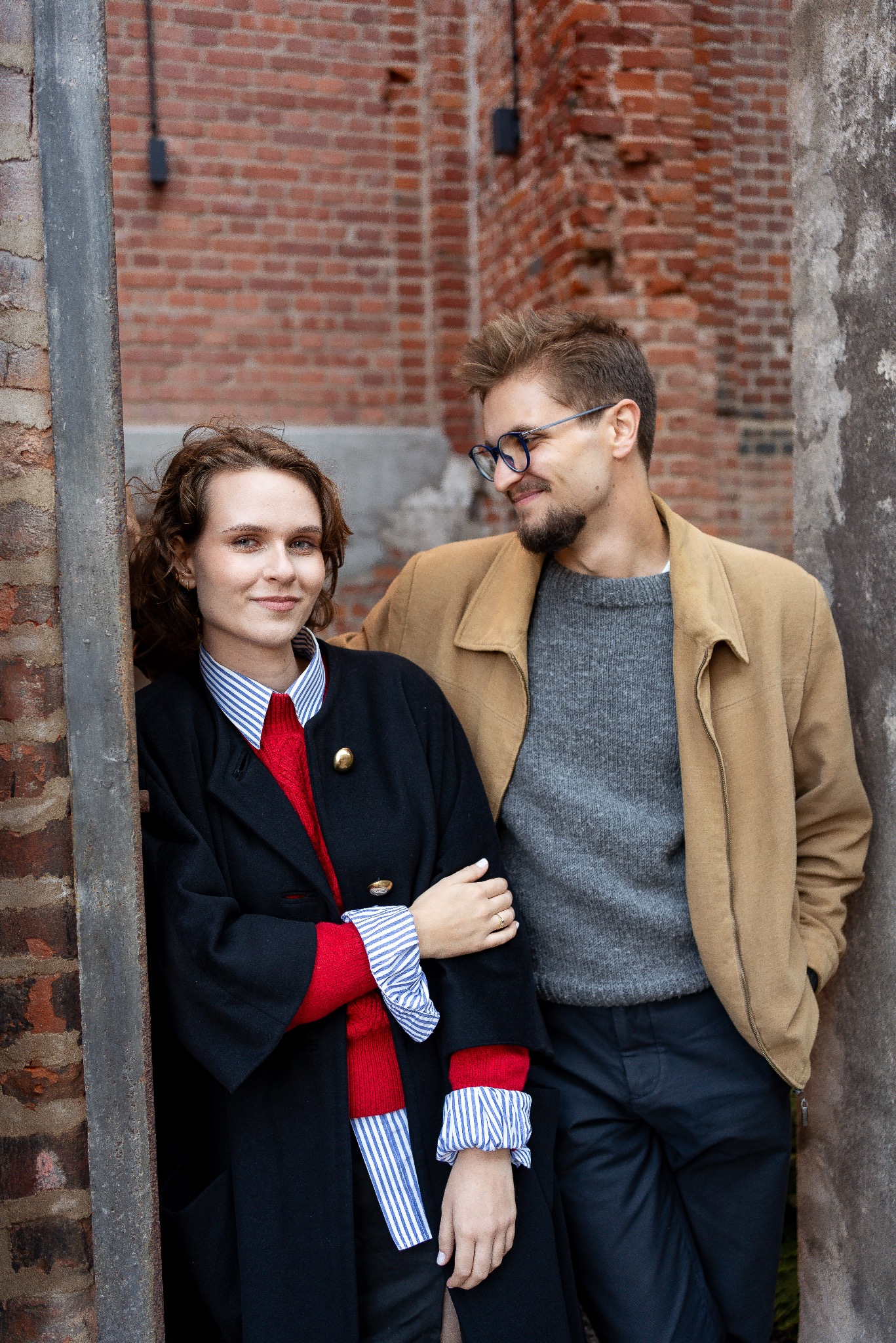 Stylowa sesja narzeczeńska: para w modnych strojach pozuje na tle ceglanej ściany i kamiennego filaru. Ona w czarnym płaszczu, on w beżowej kurtce i okularach.