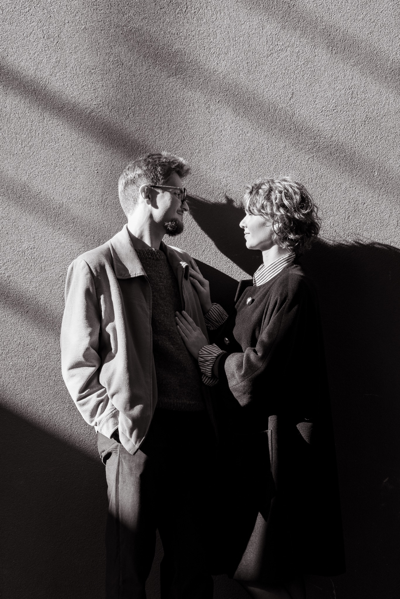 Czarno-białe zdjęcie pary, mężczyzna z brodą i okularami oraz kobieta z kręconymi włosami, stojący blisko siebie na tle ściany z cieniem, w klimacie sesji narzeczeńskiej.