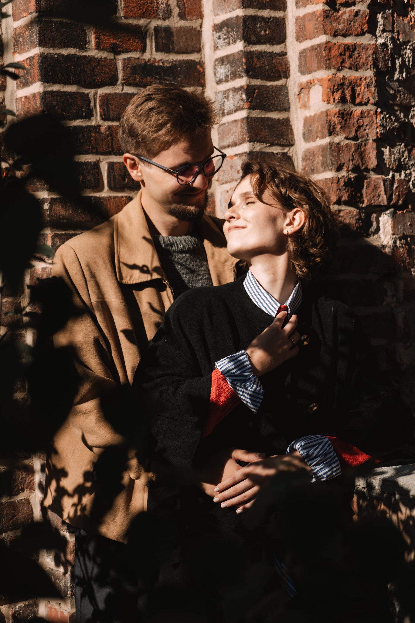 Zakochana para wtulona w siebie na tle ceglanej ściany w ciepłym, naturalnym świetle. Ciemne cienie liści dodają intymności. Sesja narzeczeńska.