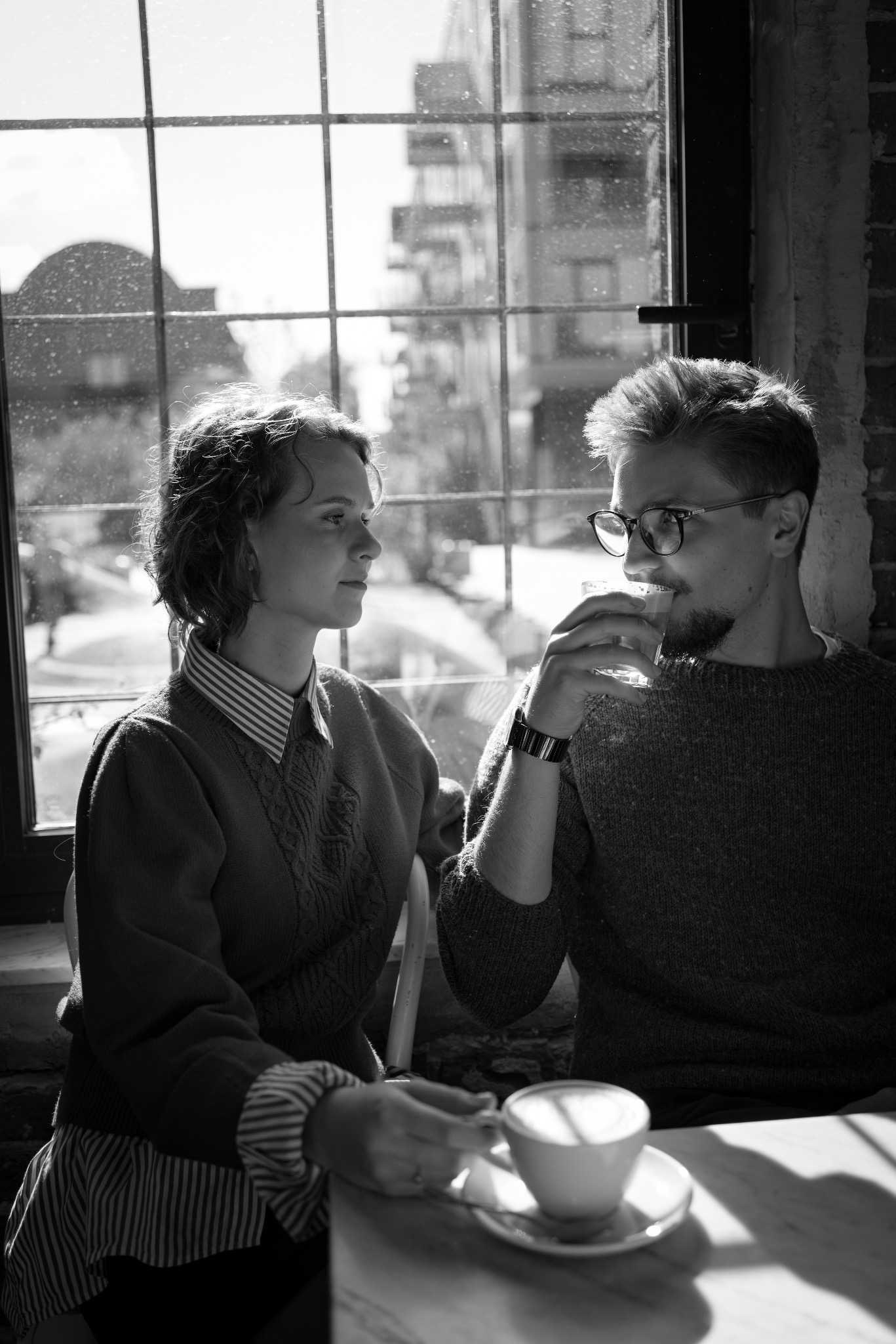 Czarno-białe zdjęcie pary siedzącej przy stole w kawiarni, z filiżanką kawy na pierwszym planie. Kobieta patrzy w bok, mężczyzna pije z szklanki. Okno w tle.