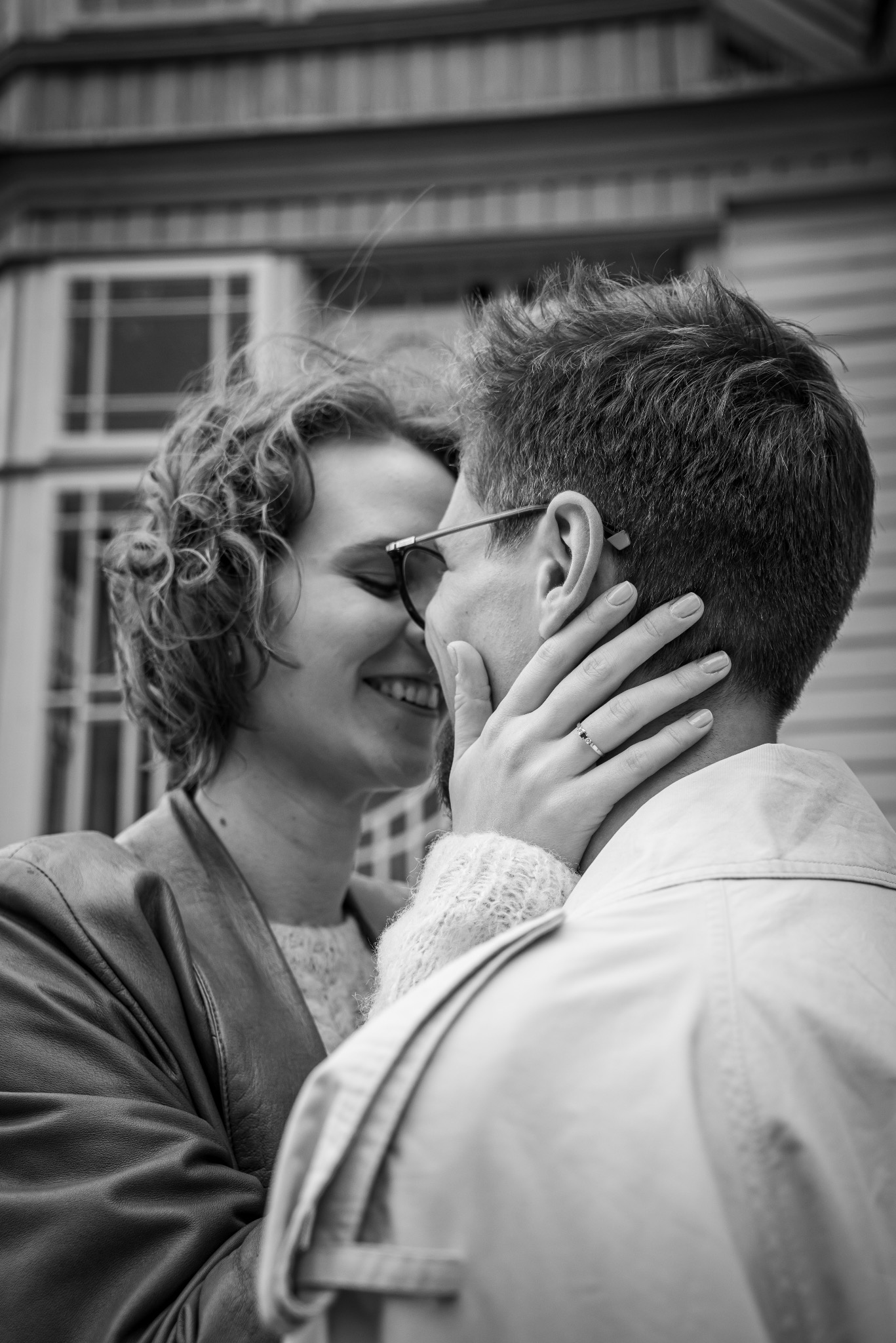 Czarno-białe zdjęcie: uśmiechnięta kobieta z kręconymi włosami dotyka twarzy mężczyzny w okularach, na palcu pierścionek, w tle budynek z oknami.