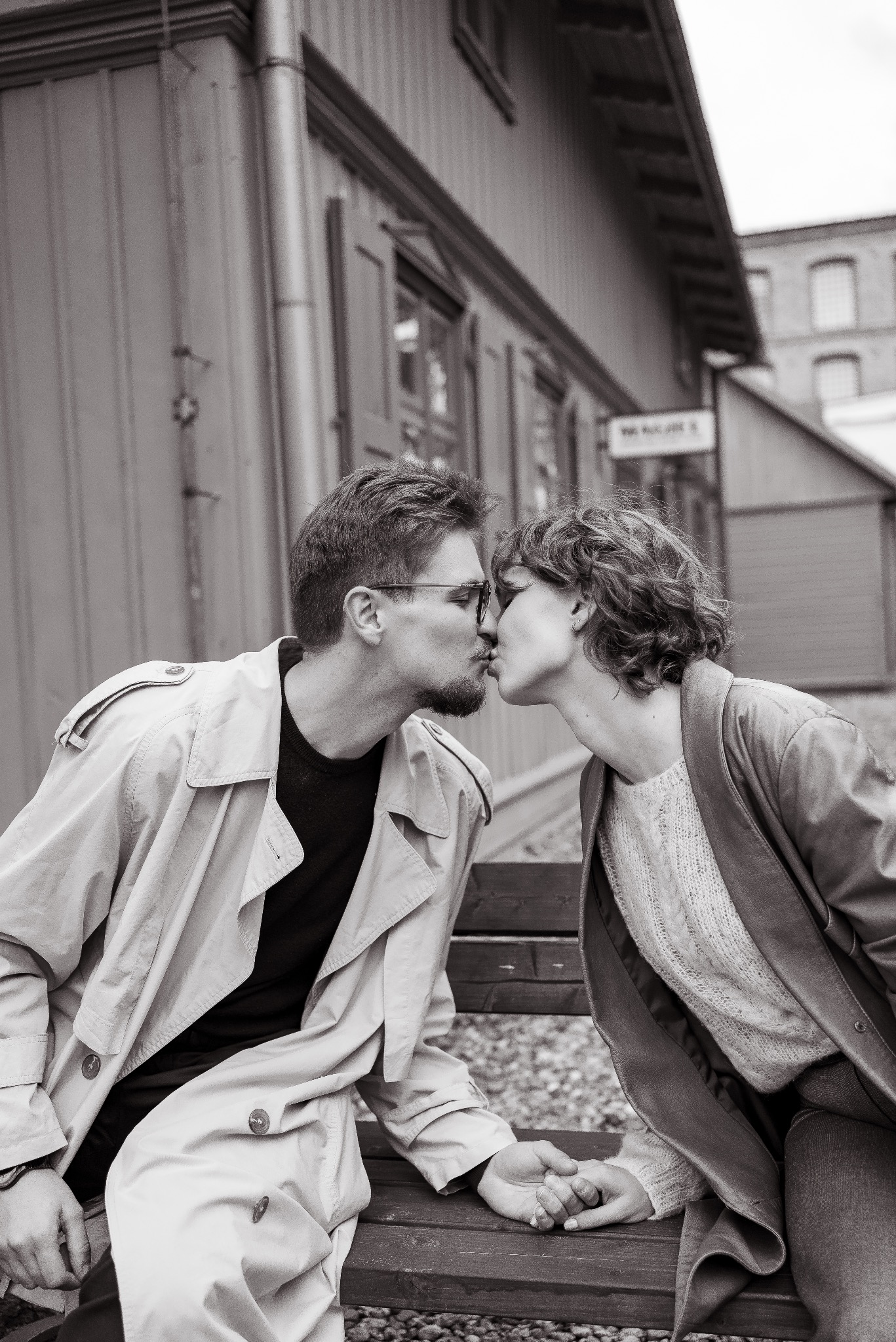 Para całująca się na ławce w plenerze, w czarno-białej tonacji. Mężczyzna w okularach i płaszczu, kobieta w kurtce. Romantyczny moment w miejskiej scenerii.