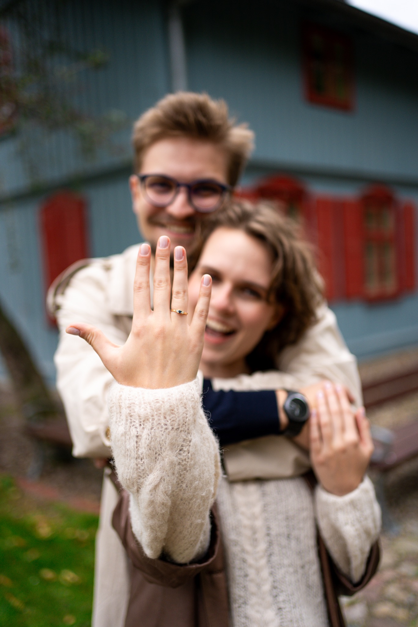Uśmiechnięta para, kobieta pokazuje pierścionek zaręczynowy na dłoni w wełnianym swetrze, mężczyzna w okularach obejmuje ją na tle budynku z czerwonymi okiennicami.
