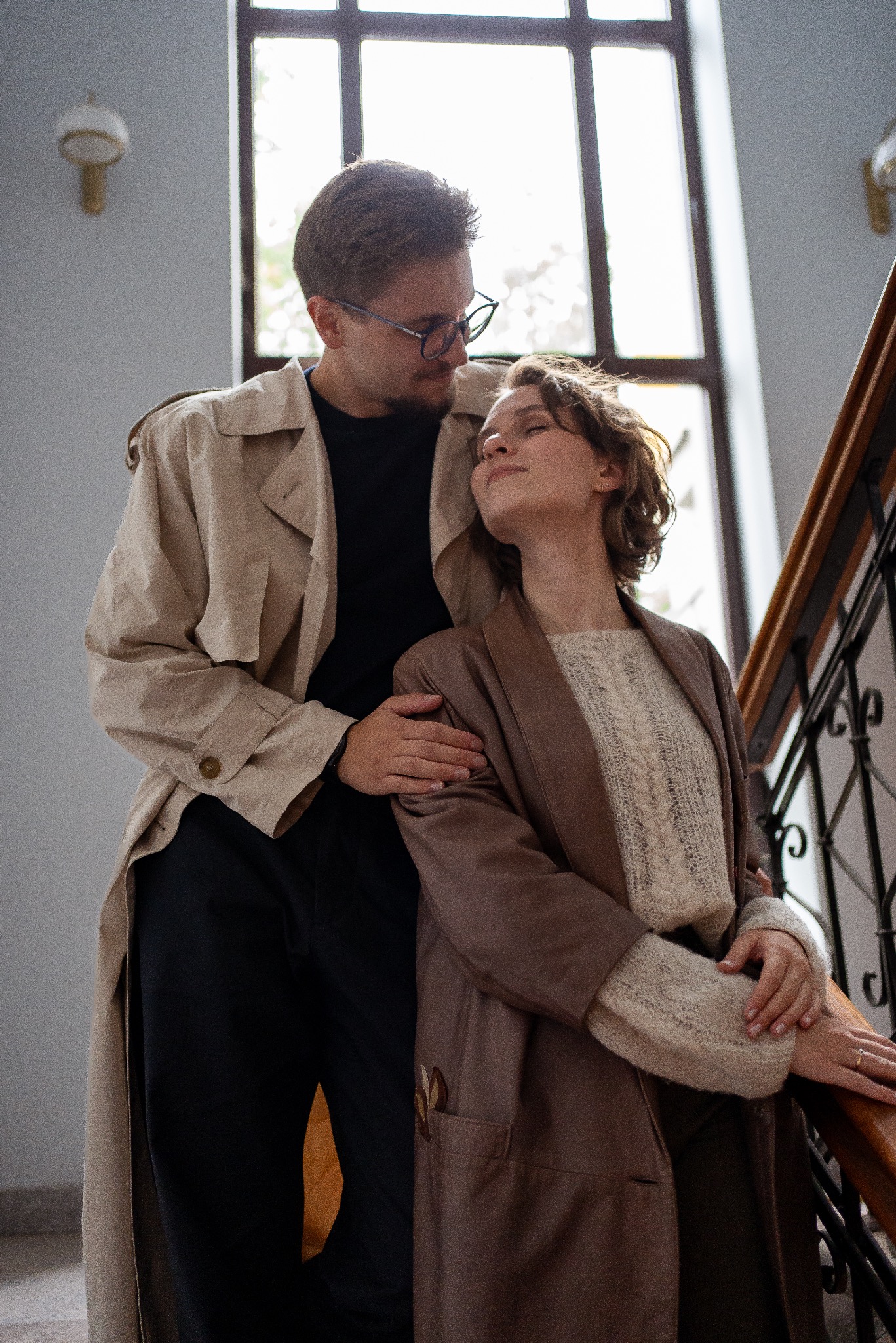 Para w jesiennych płaszczach, na tle schodów z drewnianą balustradą i dużym oknem. Mężczyzna w okularach patrzy na uśmiechniętą kobietę opartą o jego ramię.