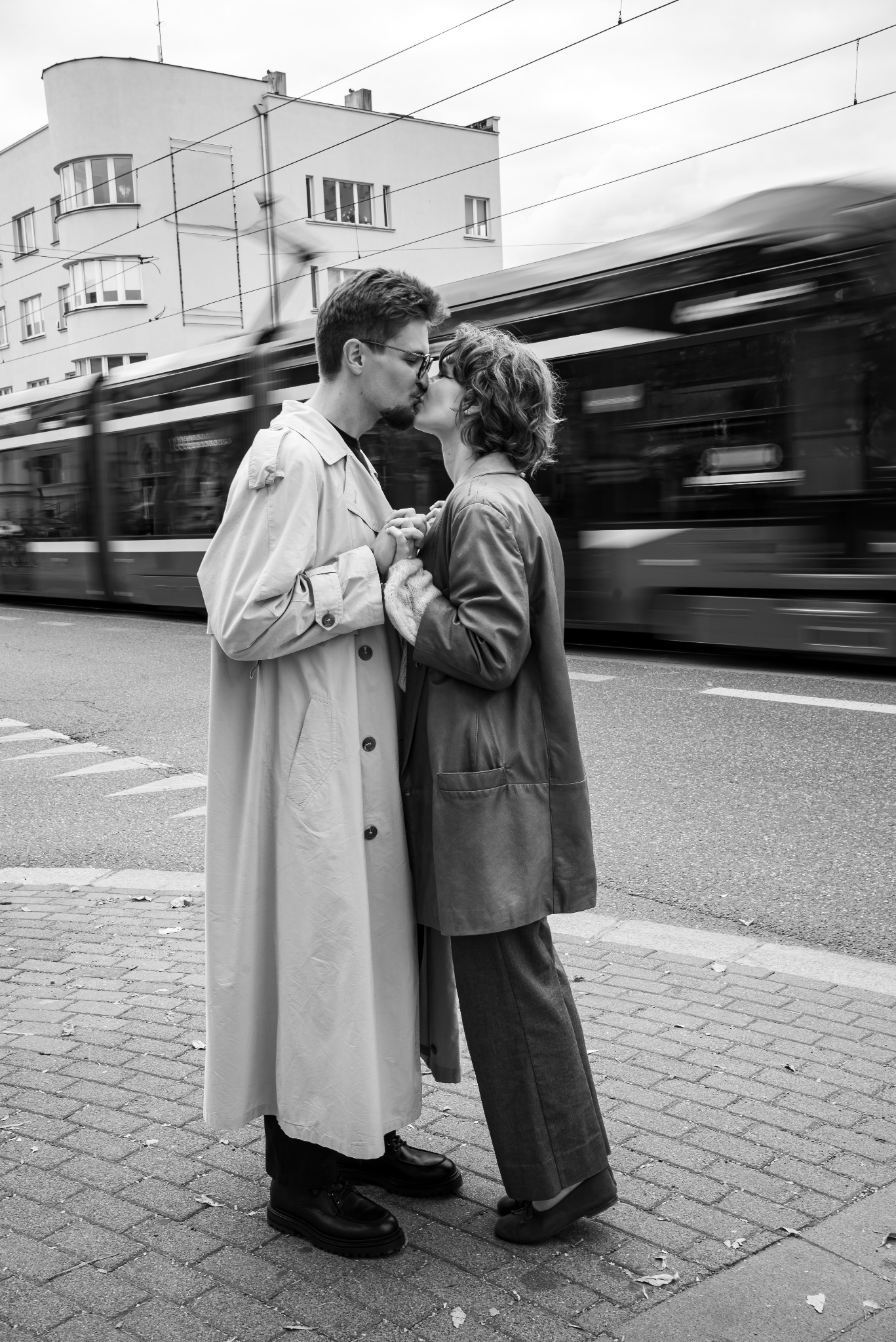 Czarno-białe zdjęcie całującej się pary na tle pędzącego tramwaju i modernistycznego budynku. Nietypowe ujęcie sesji narzeczeńskiej w ruchu.