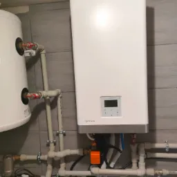 Biały bojler Aquami Series Rotenso zamontowany na szarej płytkowej ścianie, widoczne liczne rury PCV i zawory, fragment drugiego bojlera po lewej stronie.