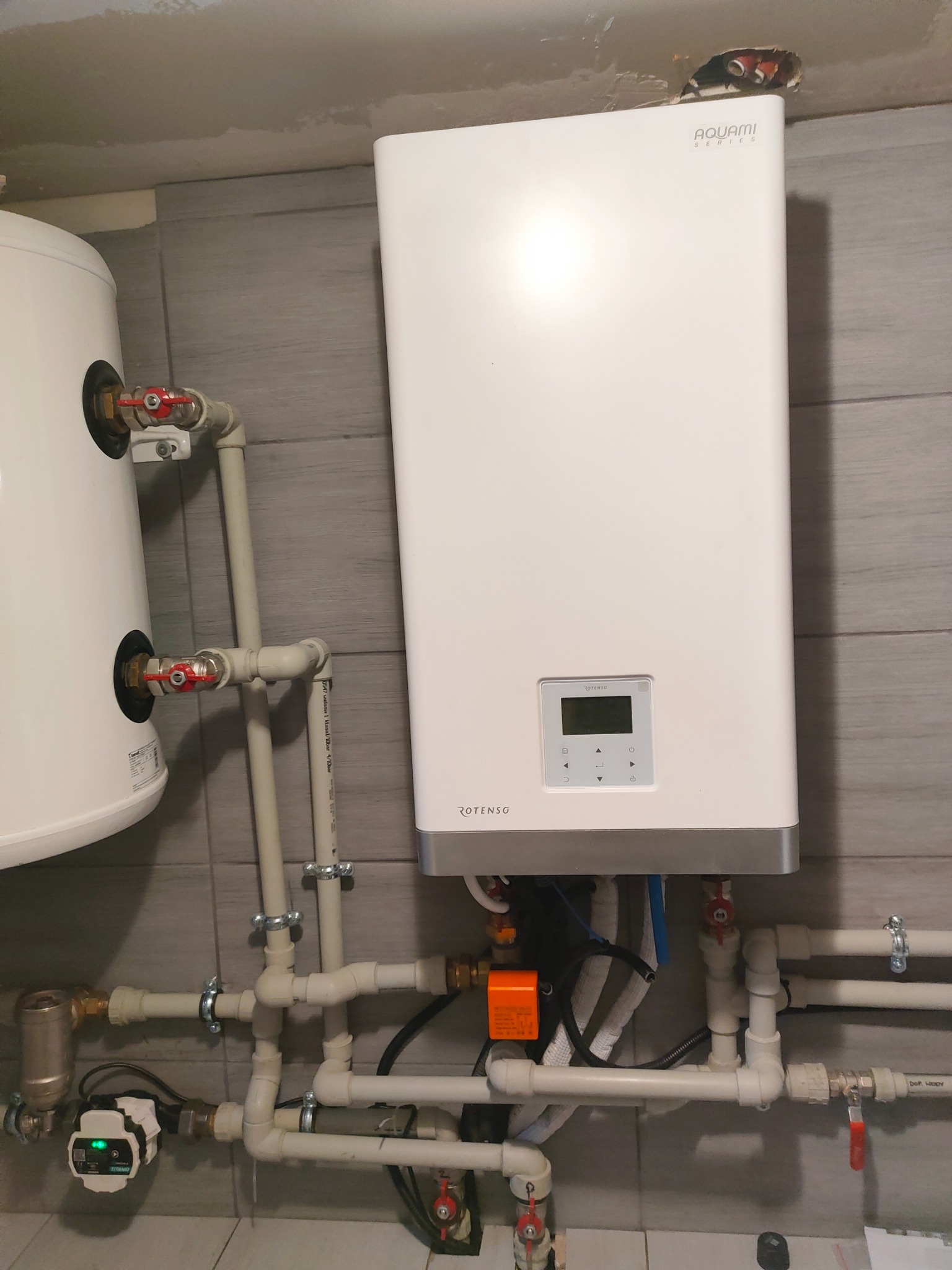 Biały bojler Aquami Series Rotenso zamontowany na szarej płytkowej ścianie, widoczne liczne rury PCV i zawory, fragment drugiego bojlera po lewej stronie.