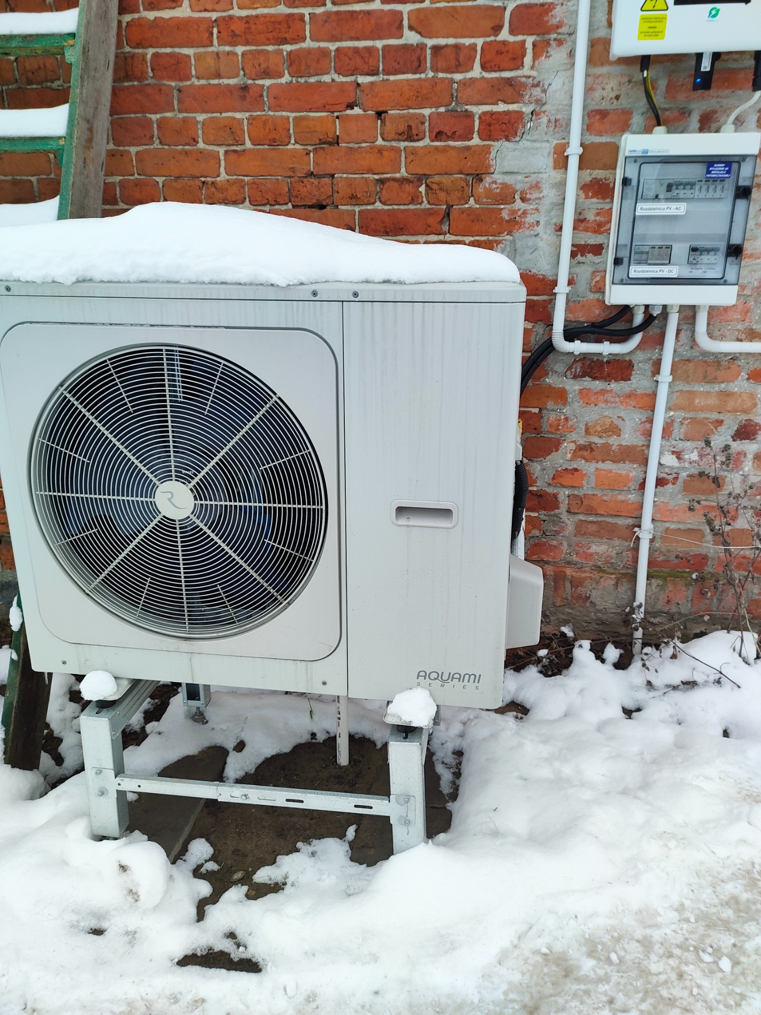 Biała pompa ciepła Aquami pokryta śniegiem, zamontowana na metalowym stelażu przy ceglanej ścianie budynku, obok skrzynka rozdzielcza PV.