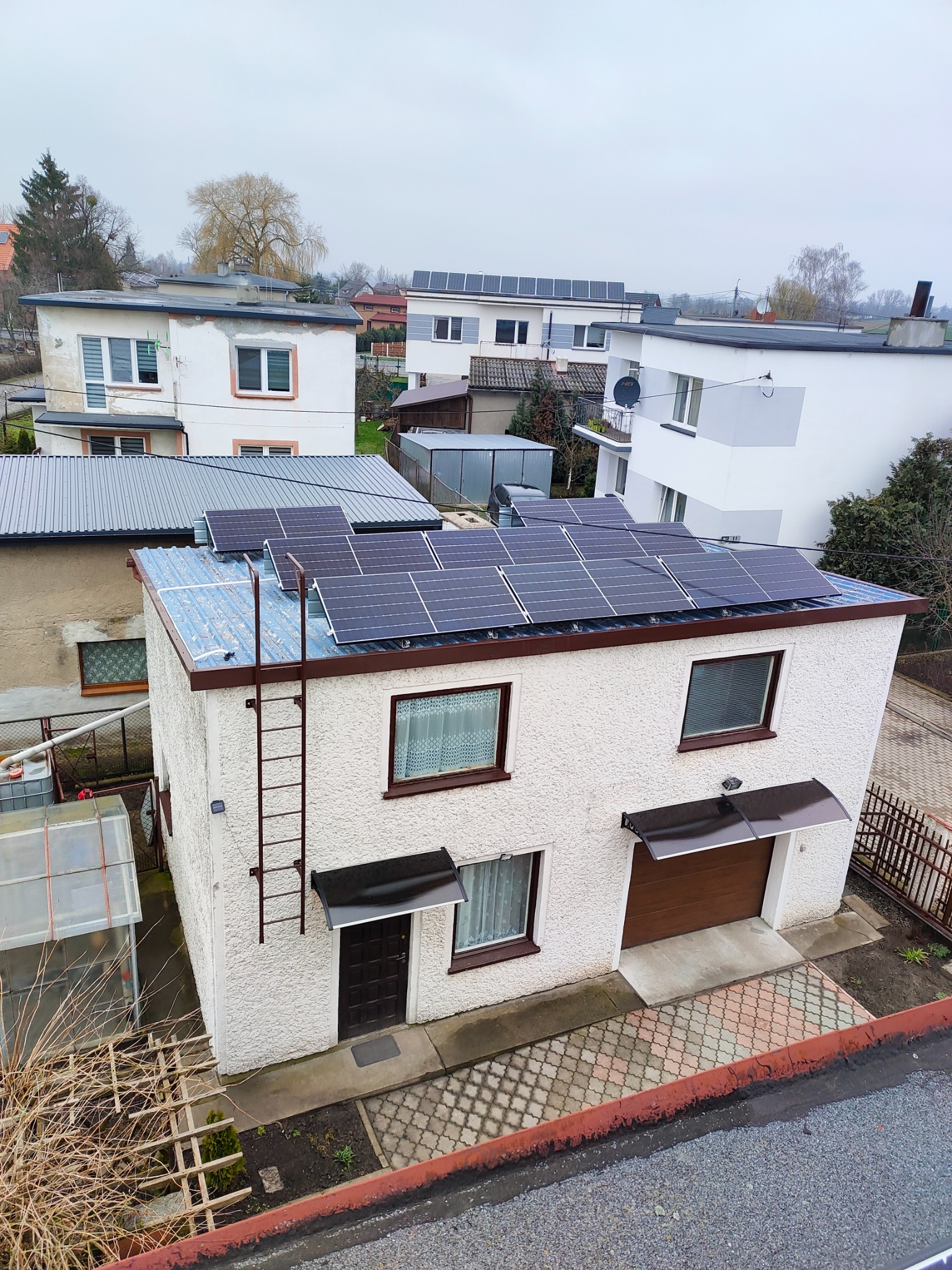 Panele słoneczne na dachu dwupiętrowego domu z fakturą baranka, drabina przymocowana do ściany, widok z góry na posesję i okoliczne budynki w pochmurny dzień.