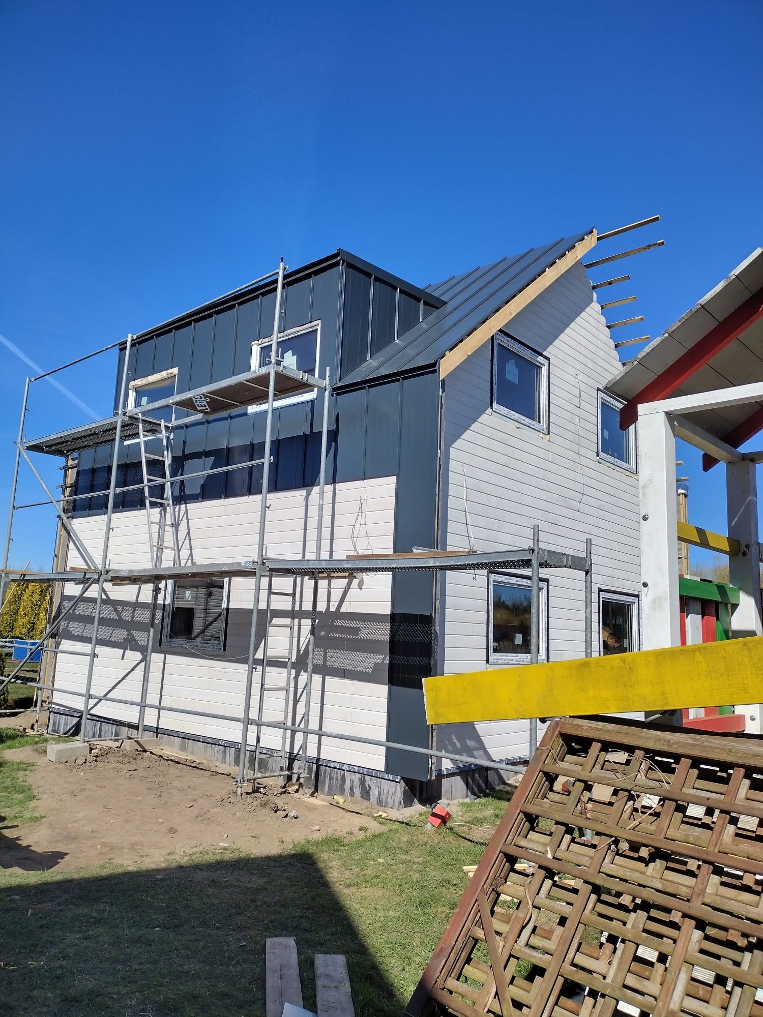 Budowa domu z szarą blachą na dachu i jasną elewacją, widoczne rusztowania i elementy konstrukcyjne. Częściowo wykończony budynek w słoneczny dzień.