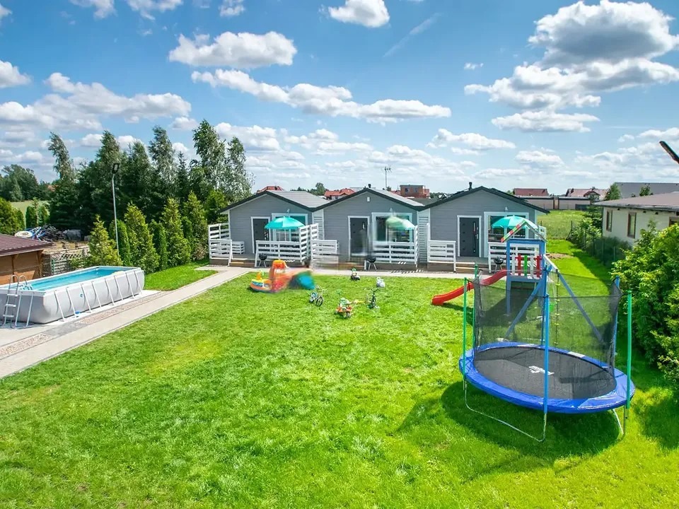 Kompleks trzech identycznych, szarych domków letniskowych z białymi balustradami, basenem i placem zabaw na zielonym trawniku w słoneczny dzień.