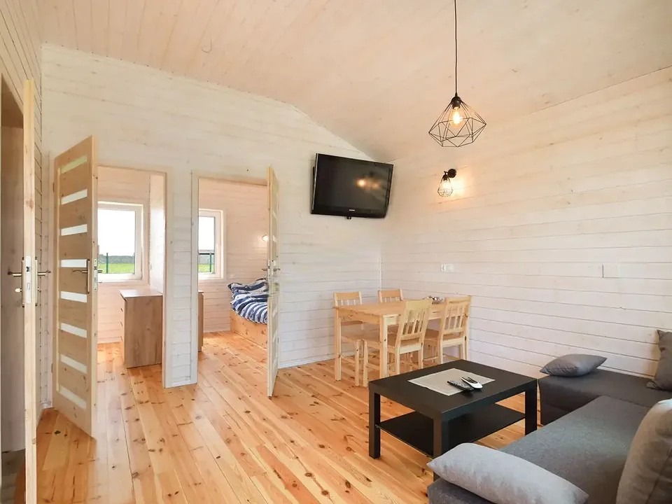 Jasne wnętrze domu z drewnianą podłogą i ścianami. Widoczny salon z szarą sofą, stół z krzesłami i telewizor na ścianie. W tle otwarte drzwi do sypialni.