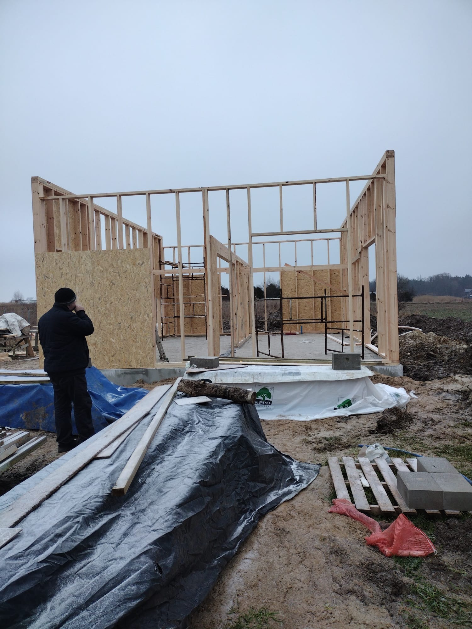 Wznoszenie konstrukcji domu szkieletowego: drewniany szkielet z płytami OSB, widoczny pracownik w czarnej kurtce, teren budowy z materiałami i narzędziami.