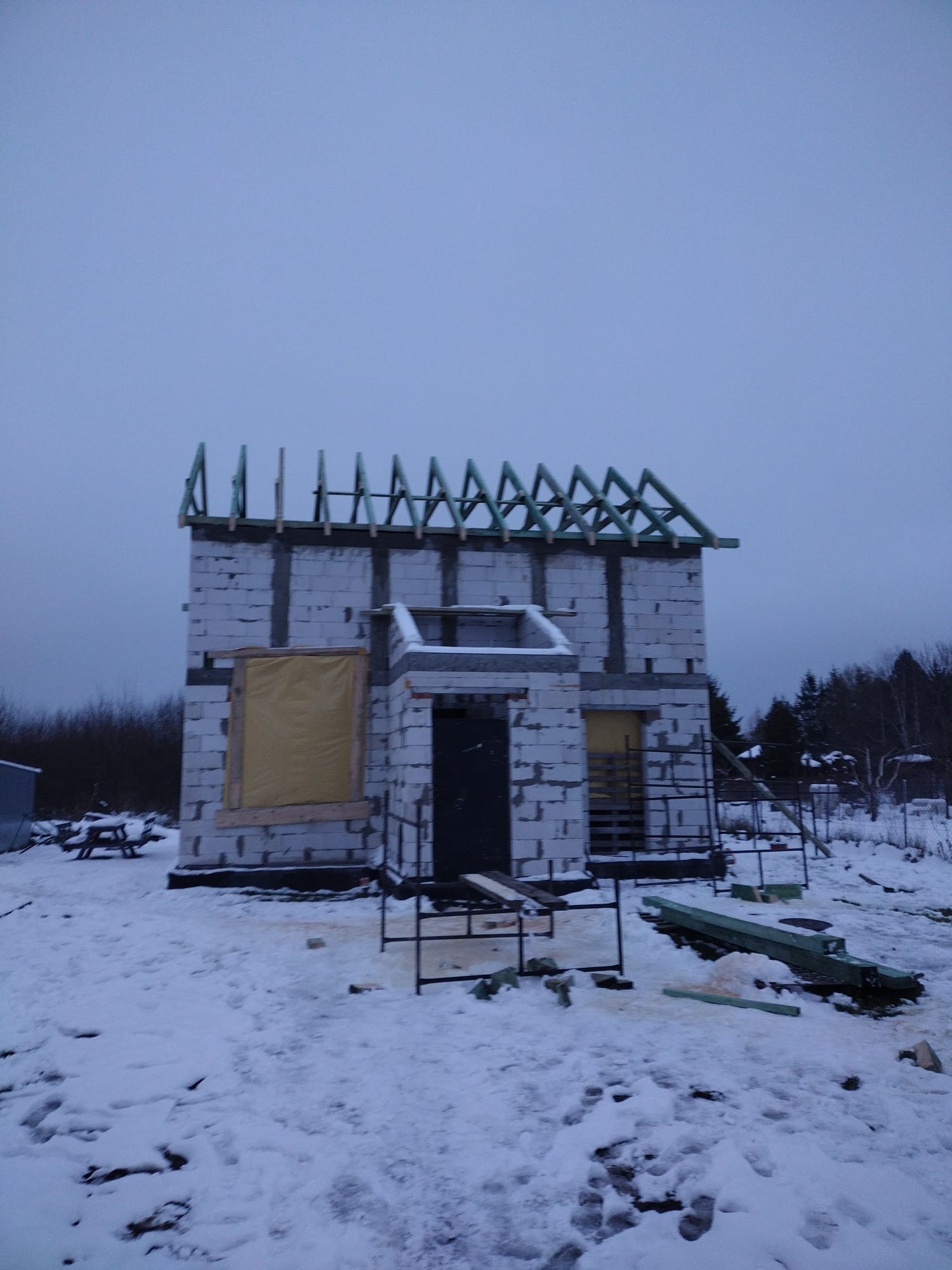 Surowy stan domu z betonu komórkowego w zimowej scenerii. Widoczne ślady śniegu, konstrukcja dachu w trakcie budowy, zabezpieczone okna.