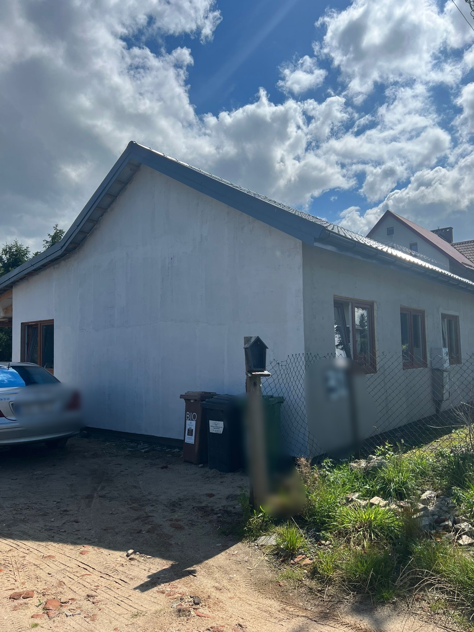 Boczny widok otynkowanego, parterowego domu z brązowymi oknami, widoczne kosze na śmieci i częściowo zasłonięty samochód. Niebo z chmurami w tle.